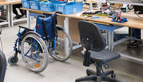 Menschen mit Behinderung - Das Bild zeigt einen Rollstuhl neben einem Bürostuhl in einer Werkstatt.